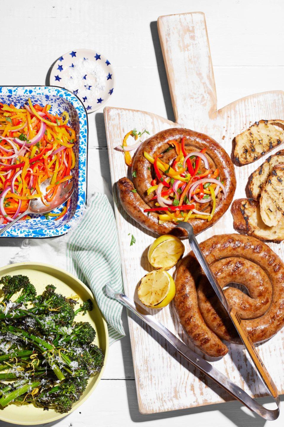 sausage pinwheels on a wooden serving board with a bowl of marinated peppers and plate of grilled broccolini to the side