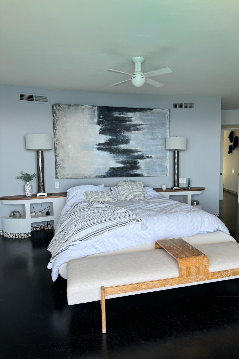 A modern hotel room with a large bed, a bench at the foot of the bed, two nightstands with lamps, and abstract artwork above the bed. Room has minimalistic decor