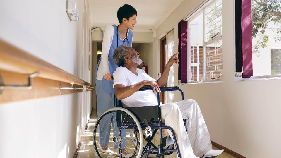 Side view of Asian female nurse and senior African-american male patient interacting with each other while standing in corridor at retirement home.