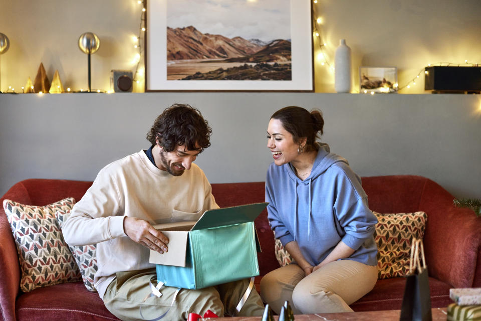 Surprised boyfriend opening gift given by girlfriend. Happy couple is celebrating Christmas at home. They are sitting on sofa.