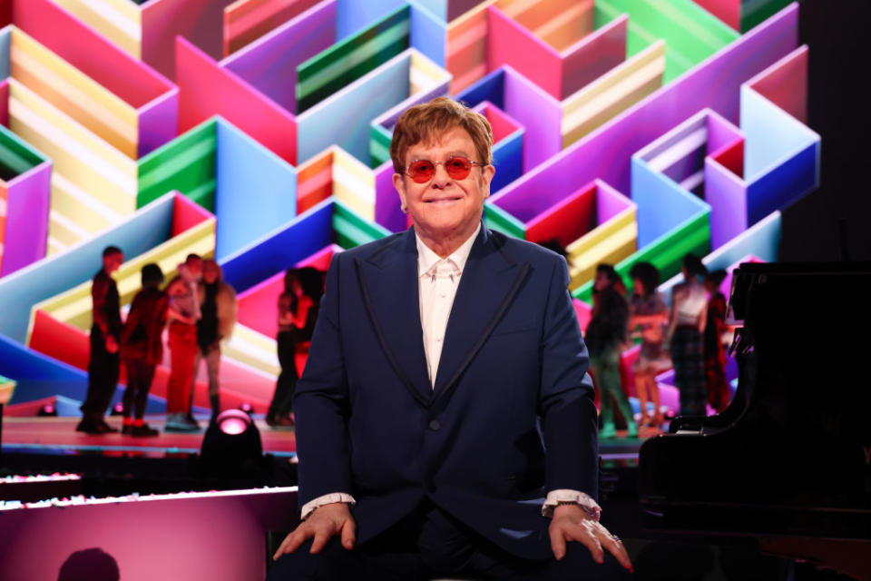 Elton John sits in front of a piano, wearing a blue suit and red-tinted glasses, with a colorful geometric background and people dancing behind him