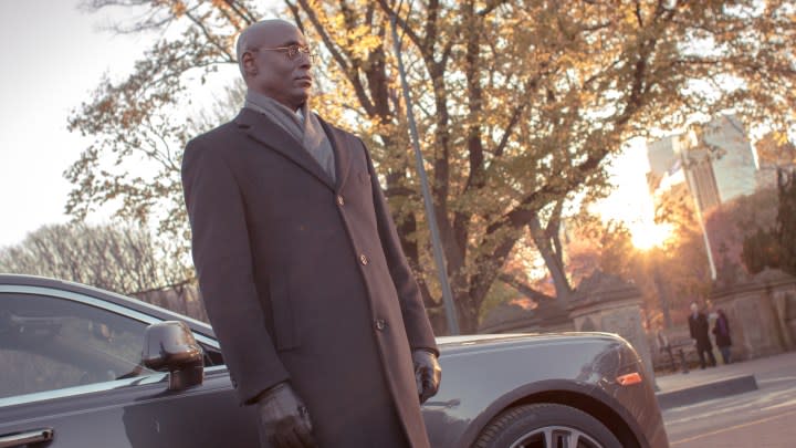 Lance Reddick stands in front of a car.
