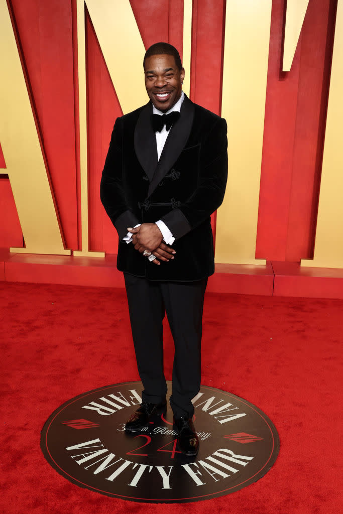 Man in a black tuxedo with bow tie, smiling, standing on a Vanity Fair emblem at an event