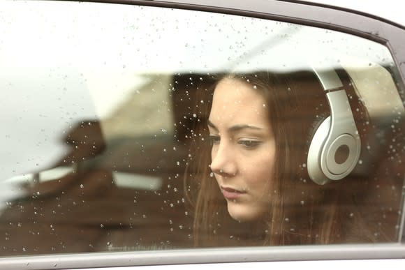 A sad woman wearing headphones, seen through a rain-washed car window.