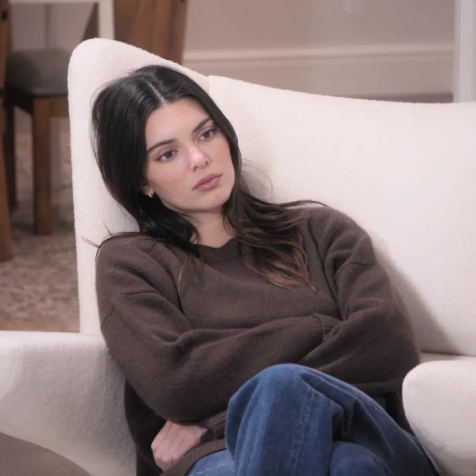 Kendall Jenner sentada en una moderna silla blanca, vestida con un suéter marrón informal y jeans azules, luciendo contemplativa.