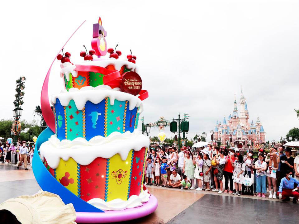 this image shows a cake installation at disneyland shanghai with crowds of people lined up behind it and a castle in the background