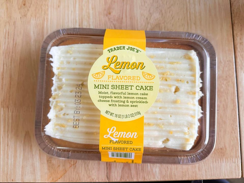 A clear package of a sheet cake with a light-yellow frosting and lemon zest on top sits on a wooden table