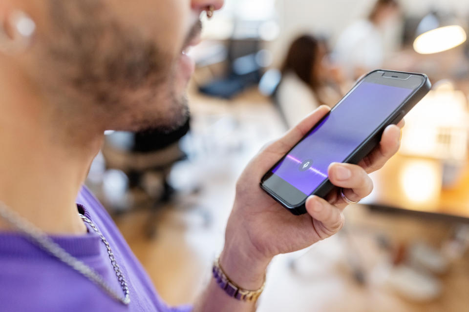 A person holding a smartphone near their mouth, seemingly using a voice assistant feature in a busy office setting with other people in the background