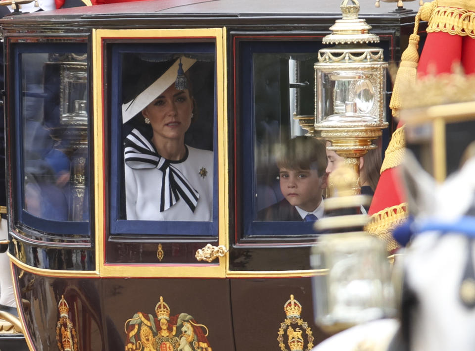 Kate Middleton and Prince Louis sit in a carriage during a royal event, both looking out the window. Kate wears a chic outfit with a large bow