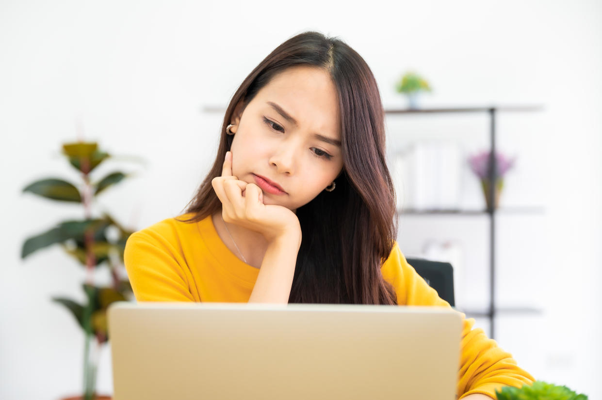 Young Asian woman in yellow looking contemplatively at her laptop, trying to remember her password