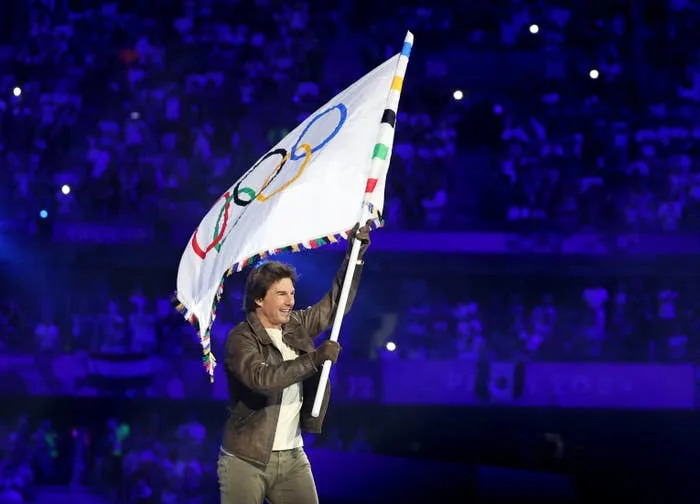 Tom Cruise waves the Olympic flag at a public event, wearing a brown leather jacket and gloves