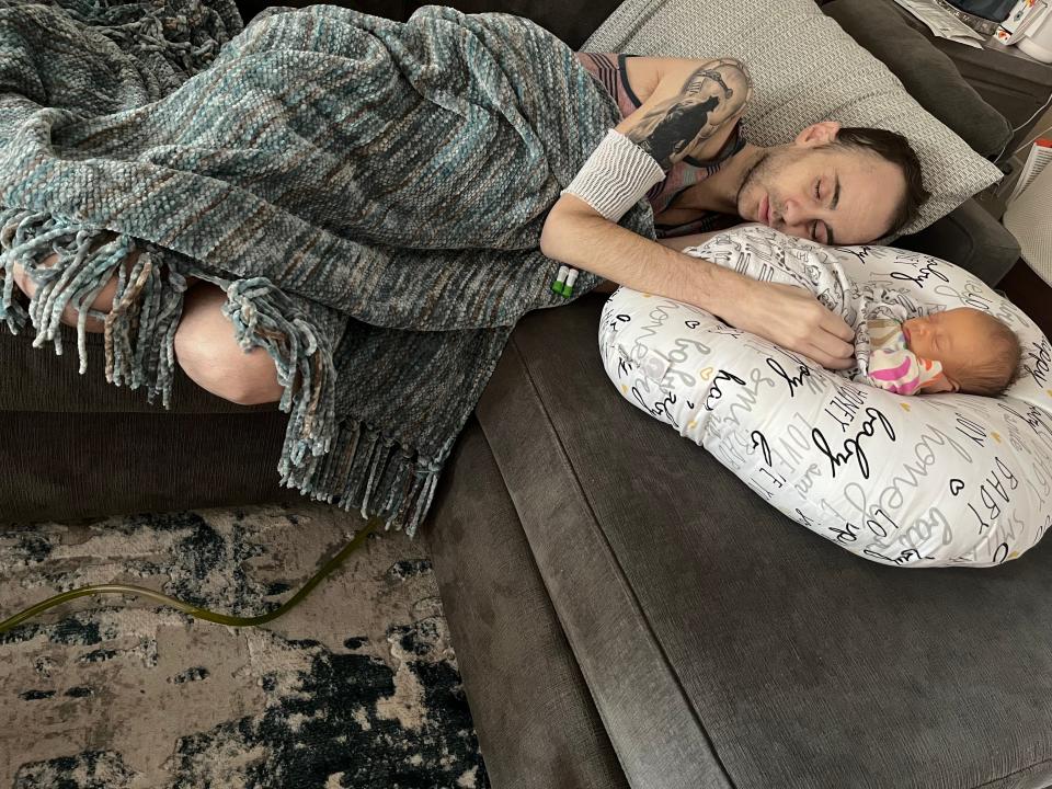 Anthony Di Laura, who has Stage IV cancer, sleeps on a couch while touching the hand of his newborn, Lila Rae, as she lies on a cushion next to him.