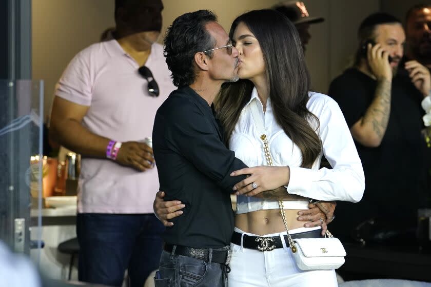 A middle-aged man holds and kisses a younger woman in a luxury box before a sporting event