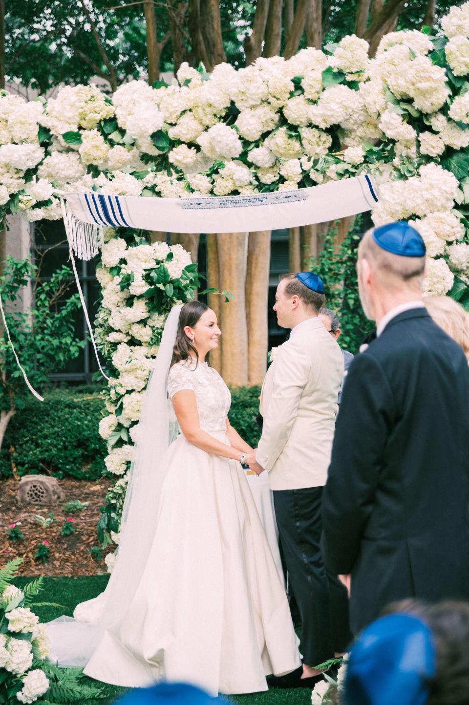 A bride smiles at her groom and holds his hands under a floral archway.