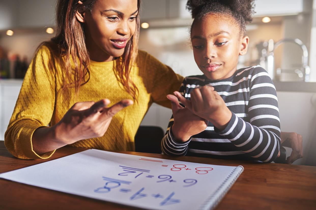 Confident young girl counting on her fingers at a wooden table with her mother sitting by her side helping her with her math homework, a notebook with simple math problems on the table and a blurred background of a kitchen
