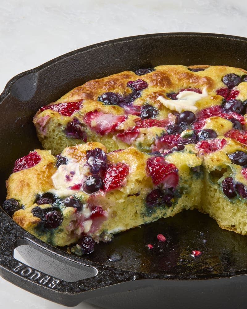 A mixed berry skillet pancake in a cast iron skillet topped with butter with some taken out and a wedge cut, ready to serve.