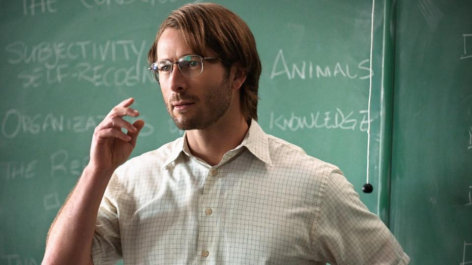 Glen Powell wearing glasses and holding his left hand up while standing in front of a chalk board.