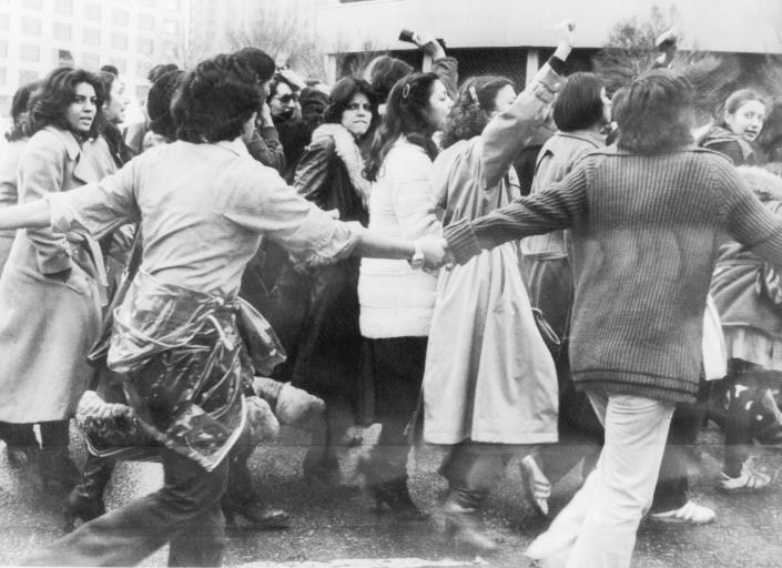 Young men wearing their hair long enough to reach their collars hold hands to form a protective ring around women raising their hands in protest.