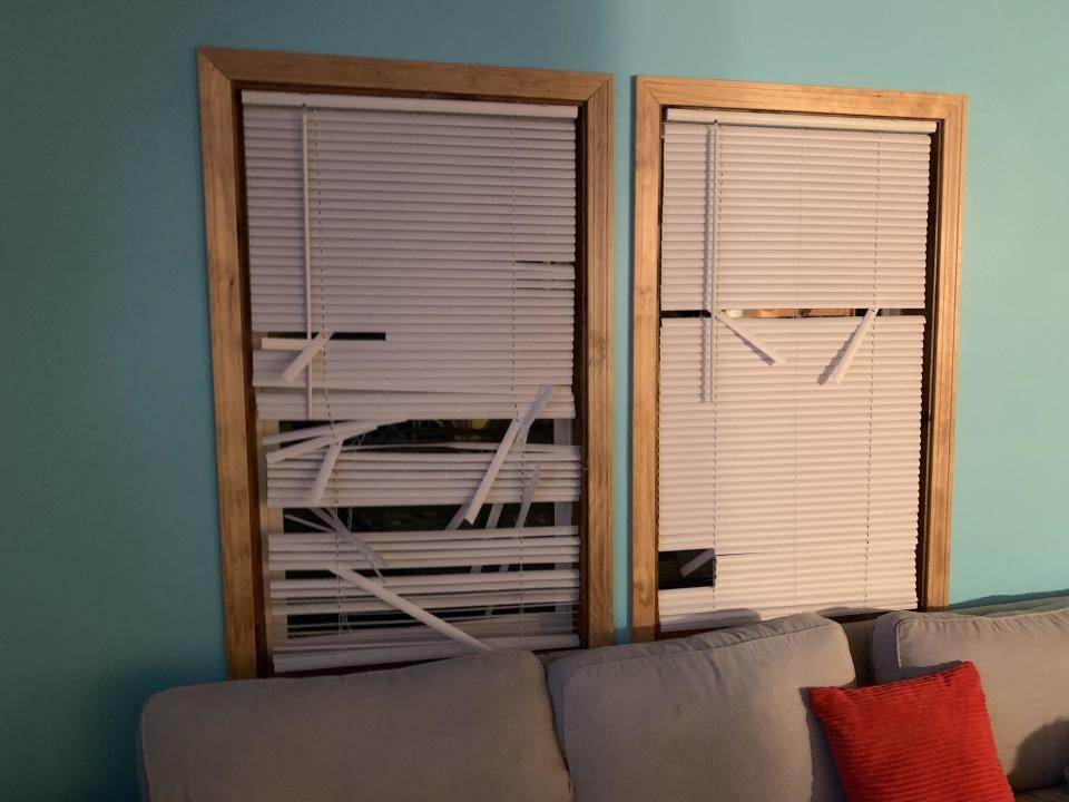 Two window frames with damaged blinds. The left blind is tangled and broken, while the right blind is mostly intact. Couch with red pillow below