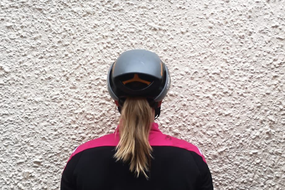 This image shows a road bike helmet from the rear being worn by someone with long hair taking advantage of the ponytail port. They are facing a white wall