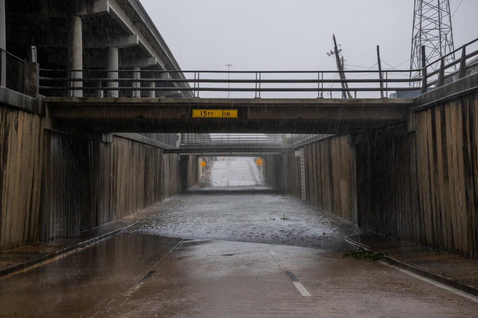 Flooded underpass with a low-clearance warning sign indicating 15 feet 1 inch. Rain pours down heavily; road is submerged. Nearby fences and trestles are visible