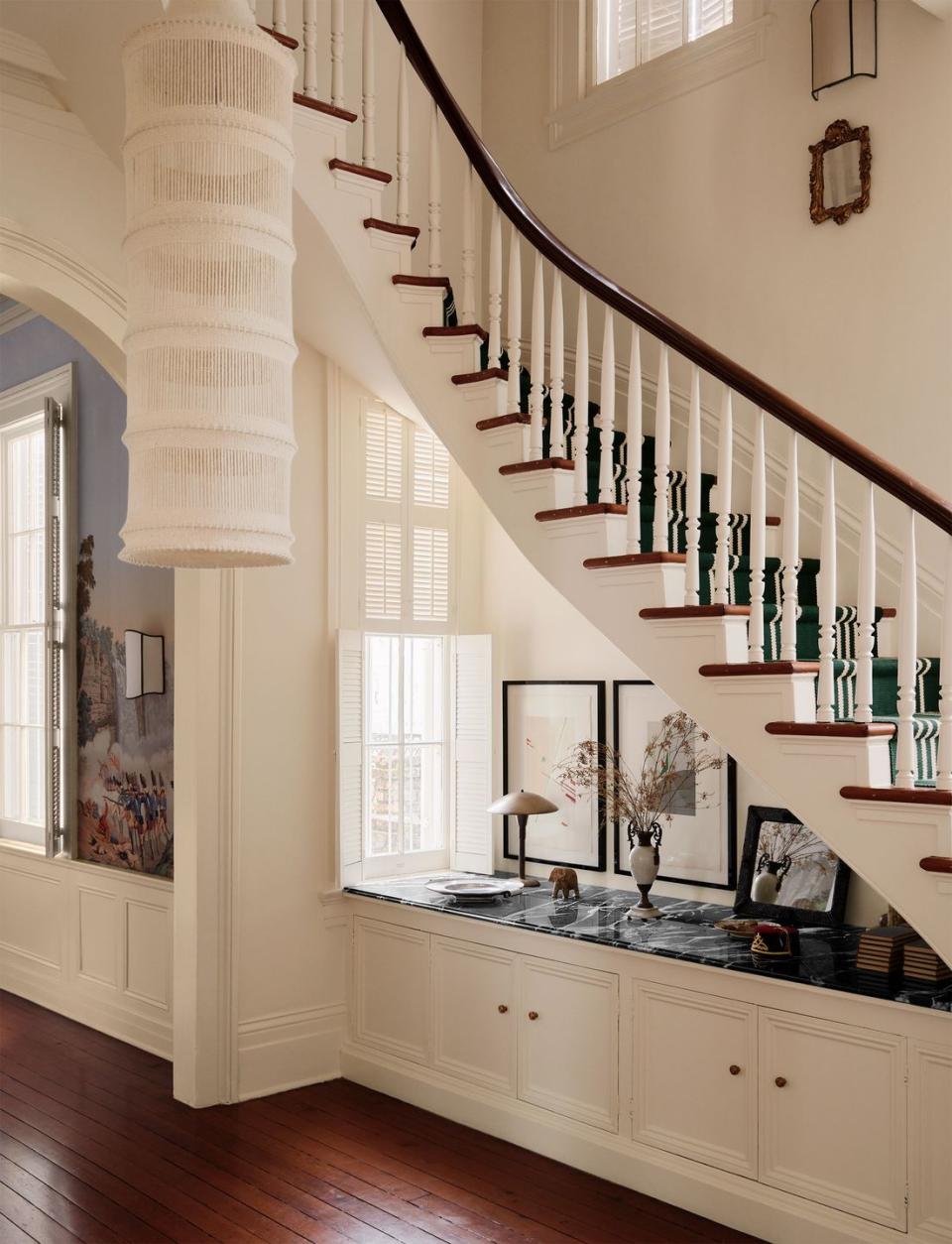 built in under a white stairway with a dark wood banister and carpeted in green with white stripes is a window and storage cabinets with a marble top that displays art objects, a macrame light fixture hangs from the ceiling