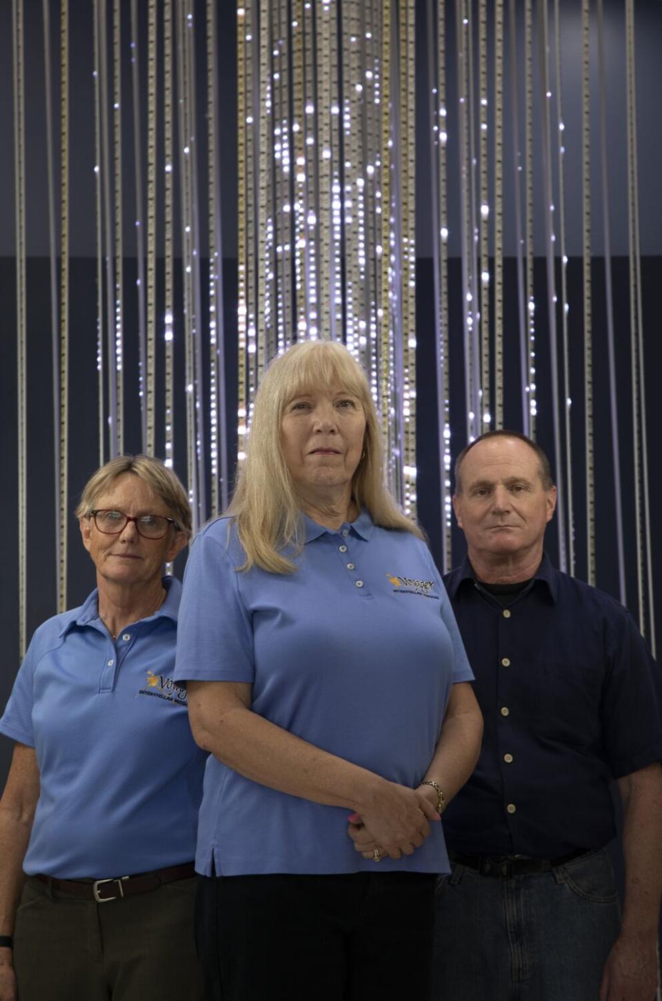 Two women in light blue shirts stand next to a man in a dark shirt