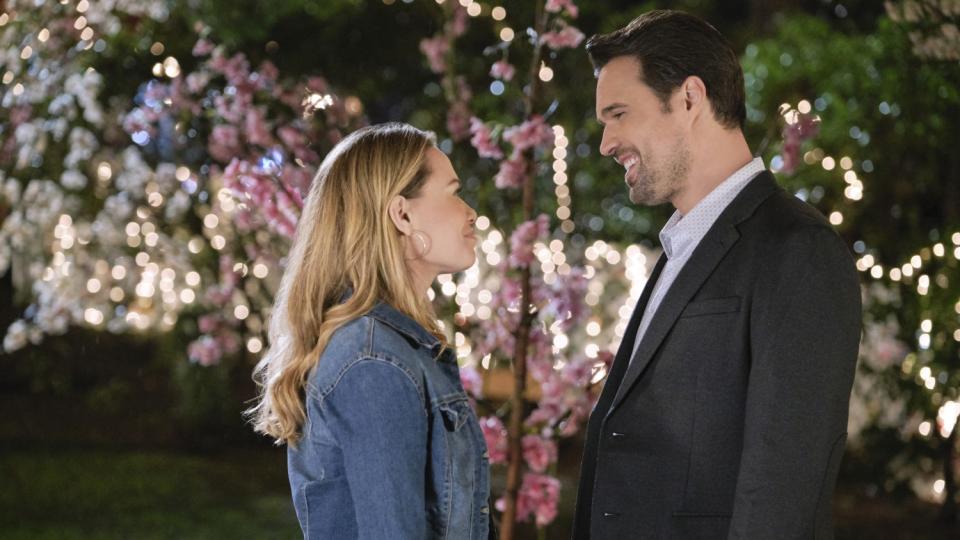 A man and a woman face each other, smiling in front of a flowering tree.