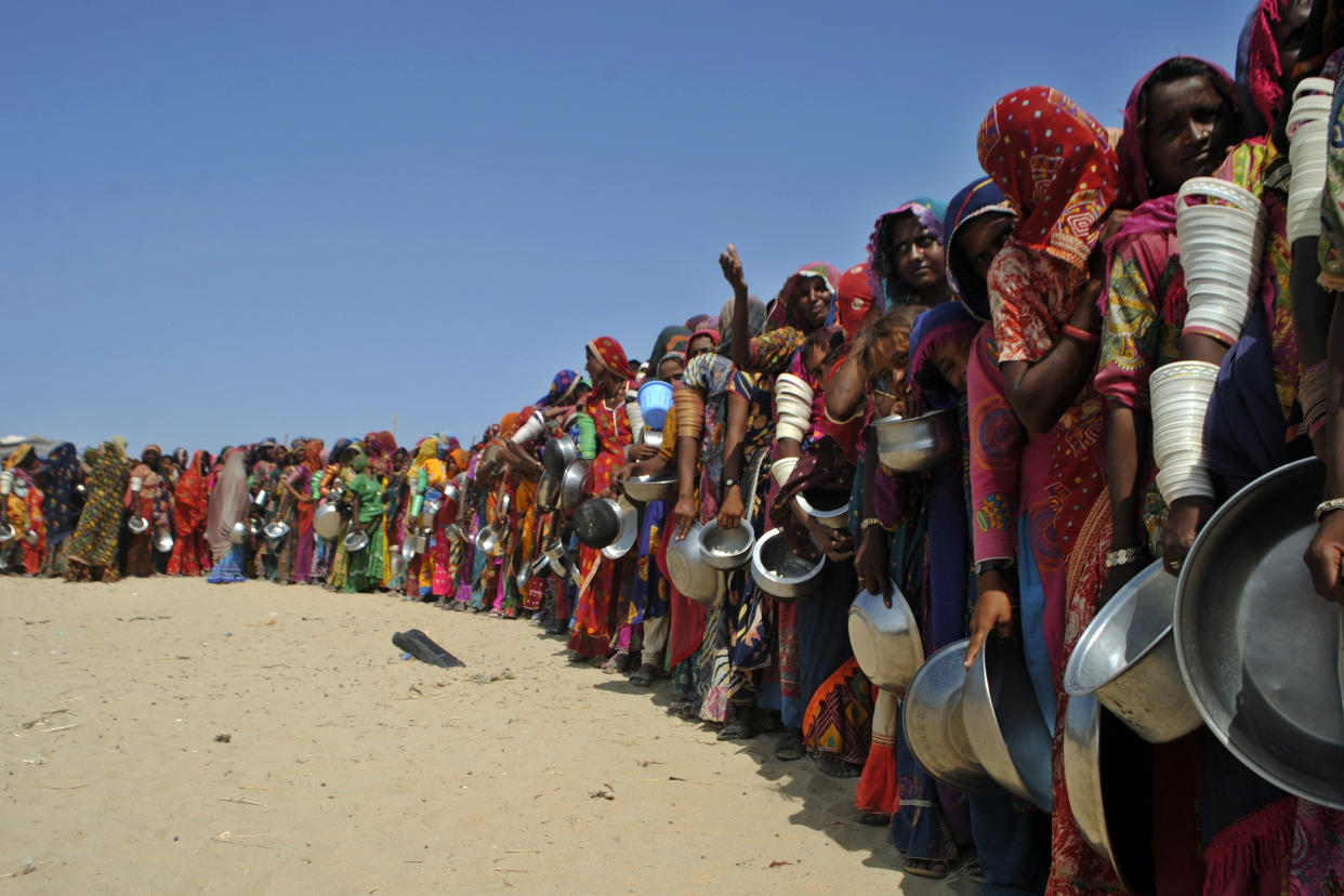 Women with colorful head coverings and bangles carry huge metal pails as they wait in a long line on desert ground.