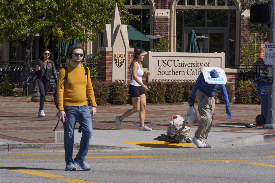 A person suffering homelessness walks past students outside the University of Southern California campus entrance in Los Angeles on Tuesday, April 16, 2024. University of Southern California officials have canceled a commencement speech by its 2024 valedictorian, a pro-Palestinian Muslim, citing "substantial risks relating to security and disruption" of the event that draws 65,000 people to campus. (AP Photo/Damian Dovarganes)