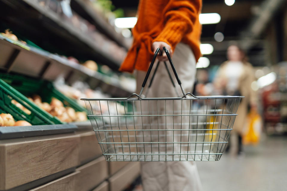 Person in a store carrying an empty shopping basket, walking by produce section