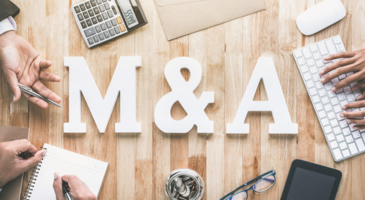 The letters "M&A" on top of a wooden table surrounded by hands, a keyboard, notepad and calculator.