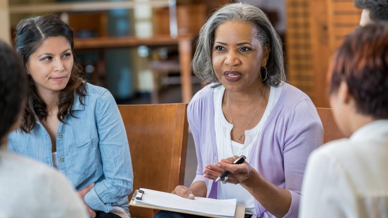 Serious senior African American female mental health profession gestures while talking with clients in a support group or group therapy session.