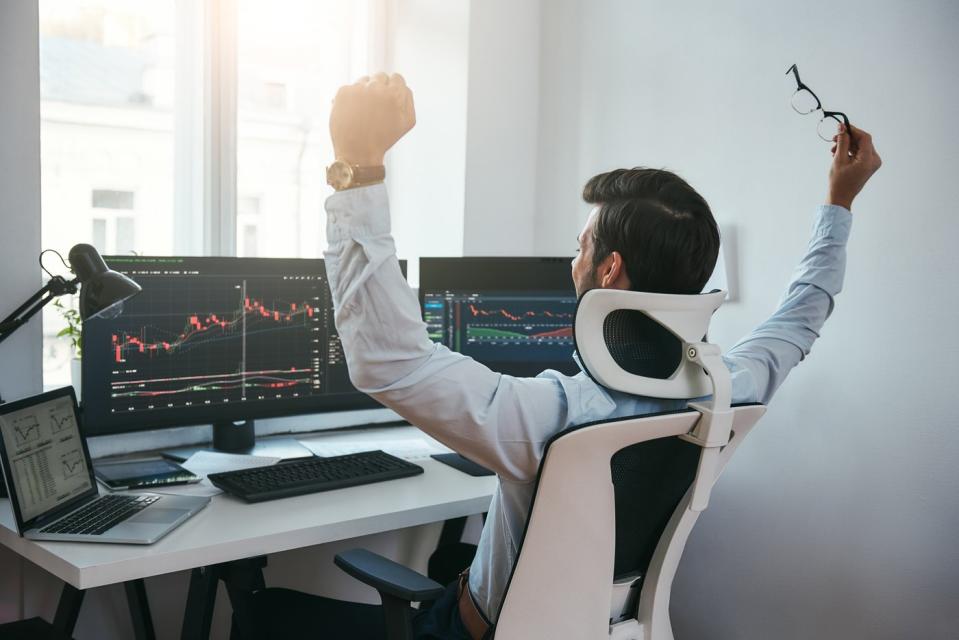 An investor raising his hands in celebration while looking at stock charts on a computer.