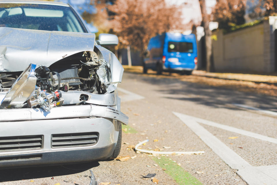 A silver car with a damaged front bumper is on a street. In the background is a parked blue van. The surroundings have light foliage