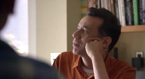 Fred Armisen sits at a desk, resting his chin on his hand, gazing thoughtfully into the distance with shelves of books in the background