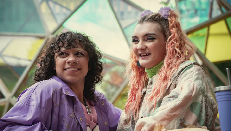 Two smiling people are dressed in pastel-colored casual wear with vibrant, curly hairstyles. They are seated together in a geometric outdoor setting