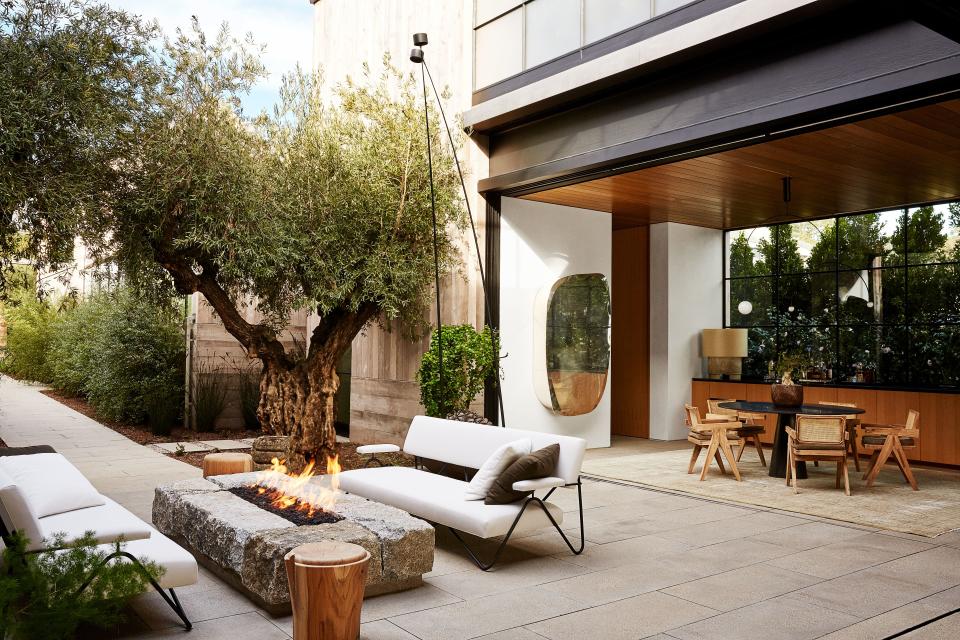 In a courtyard, a pair of sofas by William Haines Designs and teak stools flank the granite firepit. Pierre Jeanneret and Le Corbusier armchairs surround a granite table in the breakfast area.