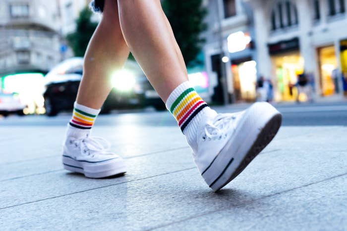 A close-up of a person walking on a city street. They are wearing white platform sneakers and rainbow-striped socks. Only the lower legs and feet are visible