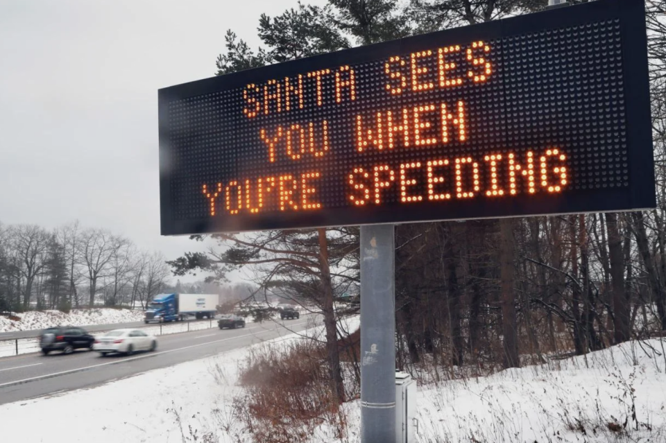 A highway message board displays "SANTA SEES YOU WHEN YOU'RE SPEEDING" beside a snowy road with cars driving by