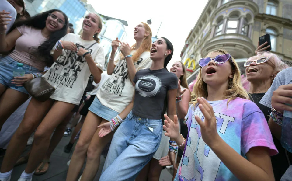 A crowd of people, some wearing Taylor Swift-branded clothing, stand and smile near several buildings.