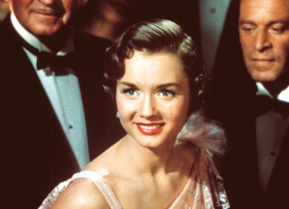 Debbie Reynolds smiles at an event in an elegant one-shoulder dress, surrounded by men in tuxedos