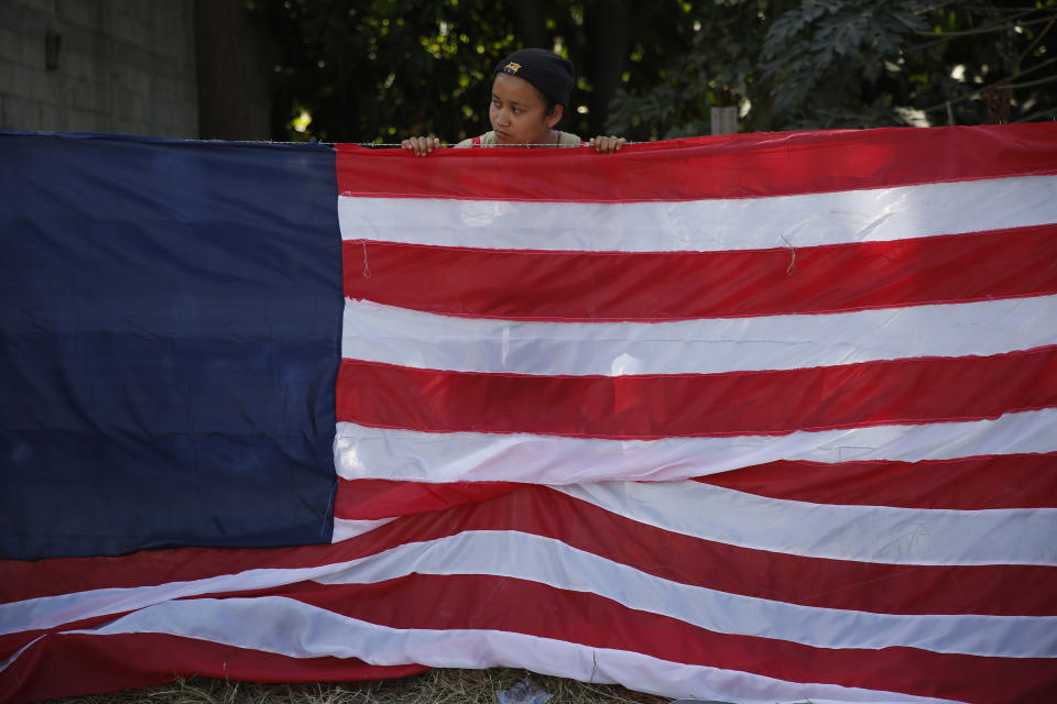 A young Central American migrant stands behind a U.S. flag made by hand by migrants, at a temporary shelter set up by authorities in Tecun Uman, Guatemala on the border with Mexico, Wednesday, Jan. 22, 2020, a location popular for migrants to cross from Guatemala to Mexico. The number of migrants stuck at the Guatemala-Mexico border continued to dwindle Wednesday as detentions and resignation ate away at what remained of the latest caravan. (AP Photo/Moises Castillo)