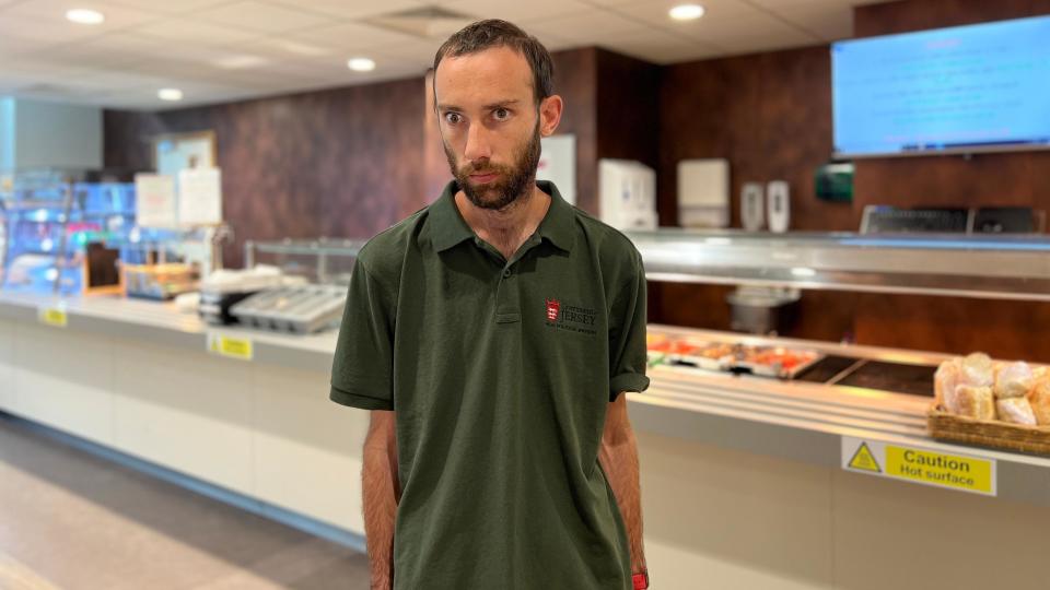 Jordan stands in front of the camera with his hands by his side. He's wearing a green T-shirt. Behind him is the food counter with a large screen to the left showing what food choices there are. A sign by the counter says caution hot surface.