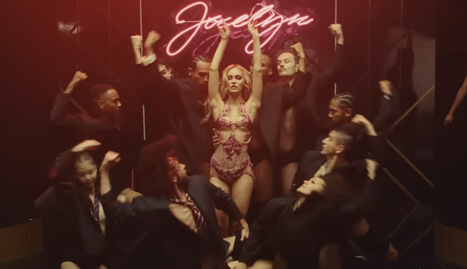 woman dancing under a neon sign reading Jocelyn with dancers in suits surrounding her