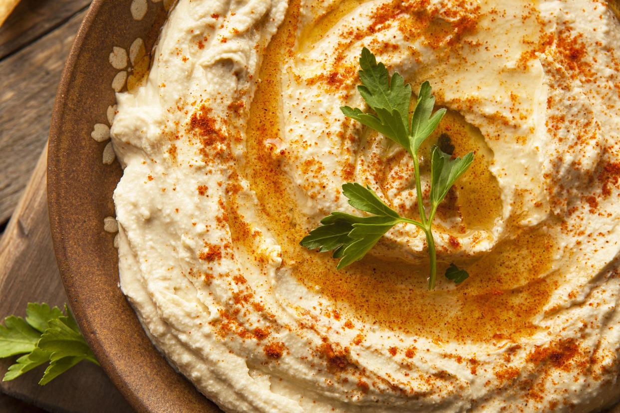 Top-view of hummus in a dark brown terracotta low-rimmed bowl, an herb leaf in the center, on a rustic wooden table on the left