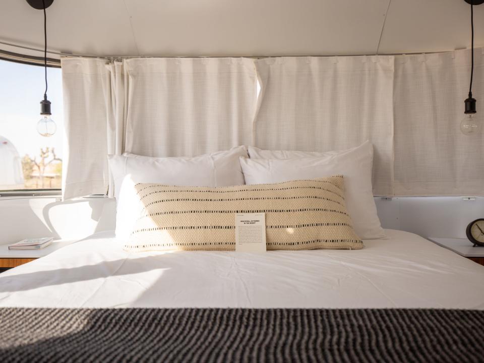 A bedroom inside Autocamp Joshua Tree's Airsteram trailer with white accents, a bed with pillows, curtains over windows.