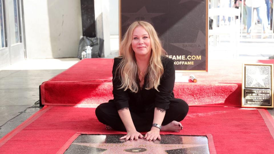 christina applegate sits on a red rug and places both her palms on top of a hollywood star, she wears all black and smiles at the camera