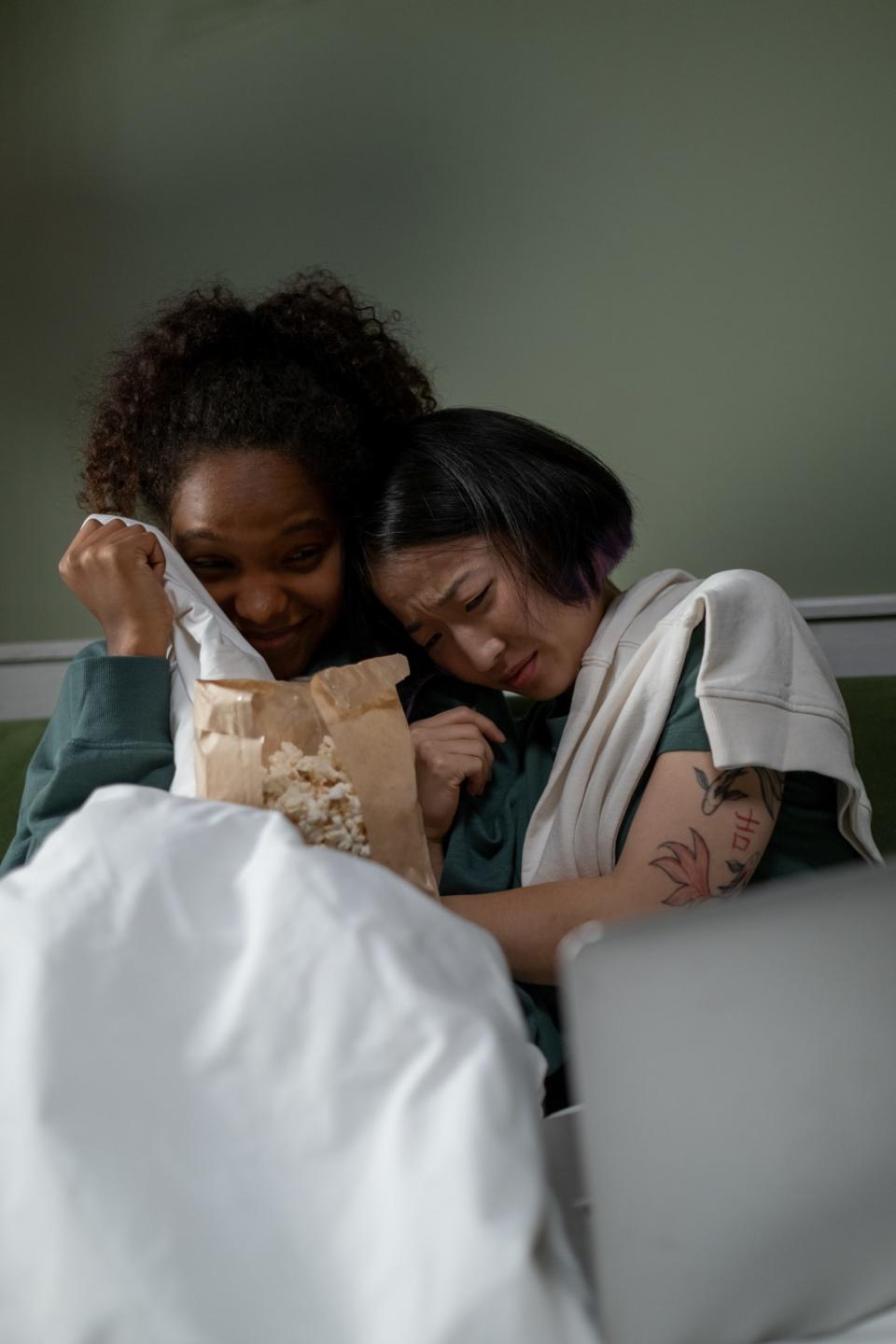 Two women hiding under a blanket while watching a horror movie