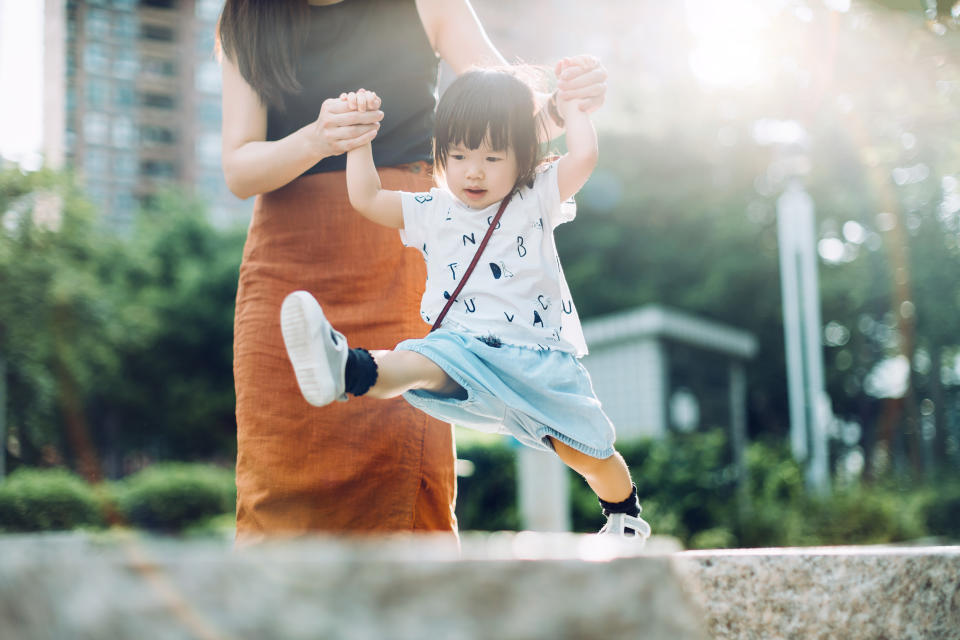 Joyful young Asian mother holding hands of little daughter supporting and assisting her walking along on stone on a lovely sunny day